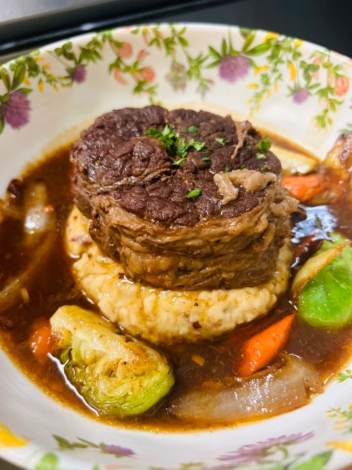 A hearty beef dish served over a bed of creamy grits with assorted vegetables in a rich sauce.