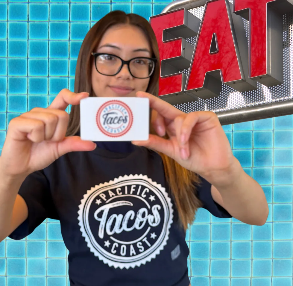 Bartender holding Pacific Coast tacos Gift Card wearing a Pacific Coast tacos T-shirt standing in front of an aquaa blue wall.