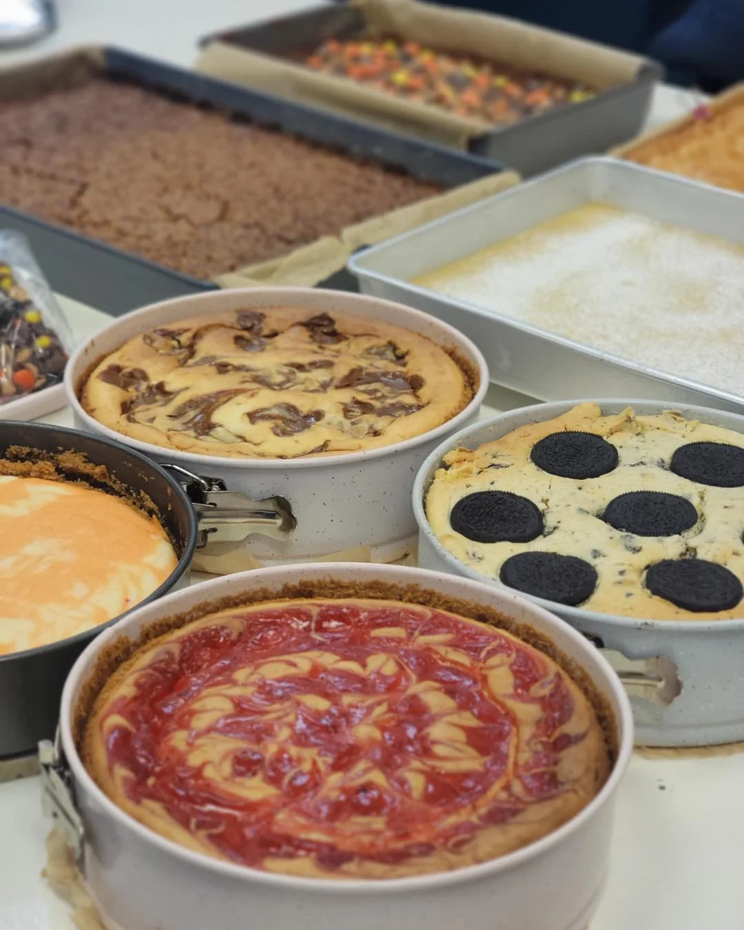 Assorted cheesecakes and baked desserts displayed on a table. The front cakes include a strawberry swirl cheesecake, an Oreo cheesecake topped with whole cookies, a marbled cheesecake, and a plain cheesecake. Large trays of brownies and other baked bars are in the background.
