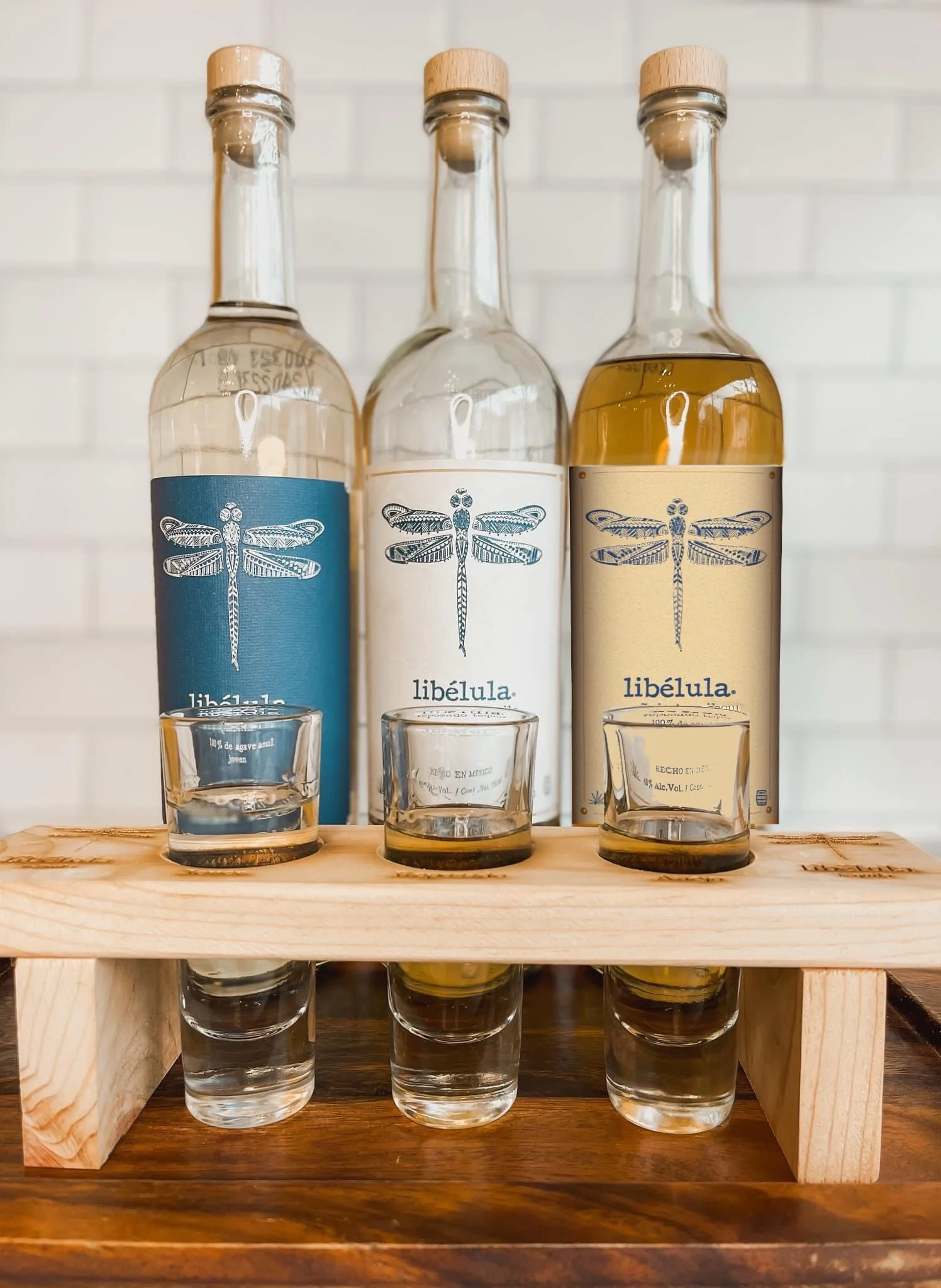 Three bottles of libélula mezcal with tasting glasses on a wooden stand.