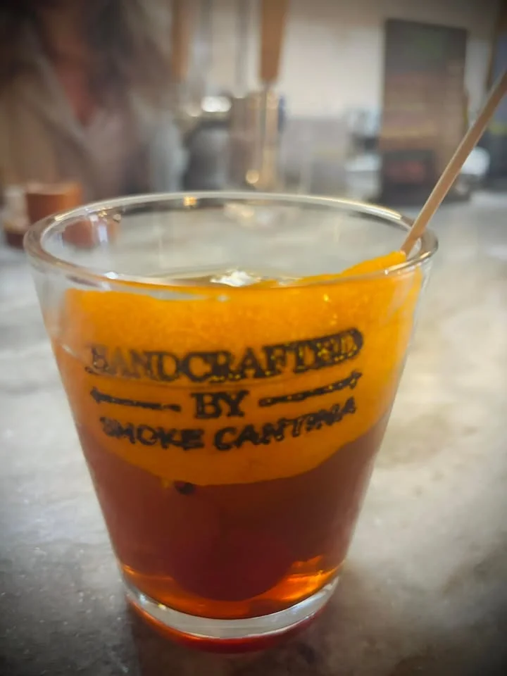 A glass with a rich amber drink, garnished with an orange peel, displayed against a blurred background.