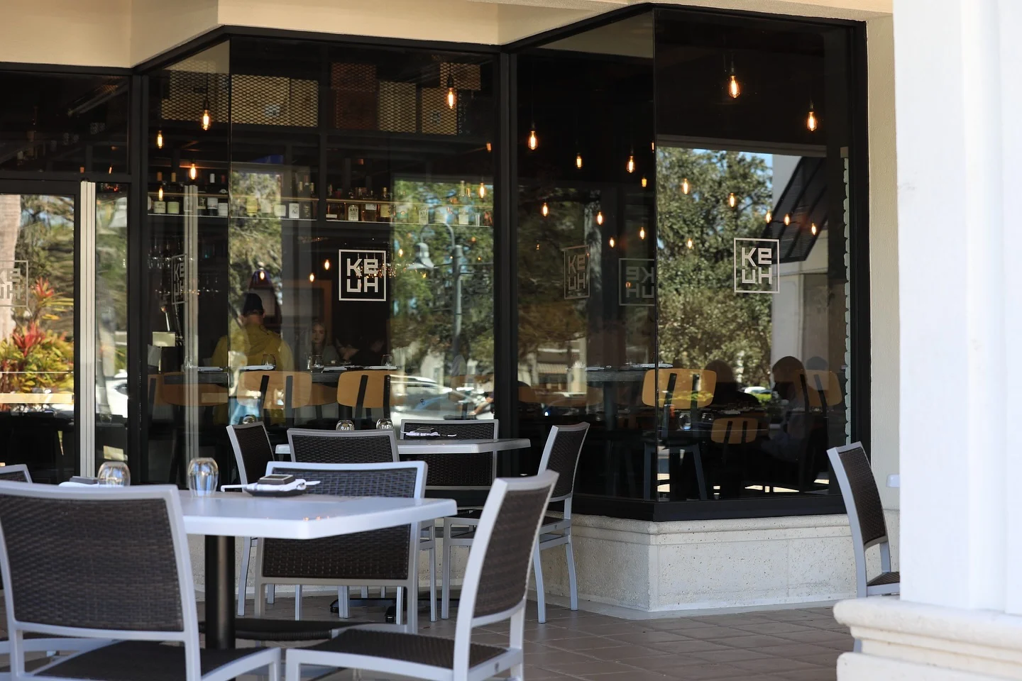Ke-uH Boca Raton patio featuring dining tables and chairs set up next to a large corner glass window that displays the Ke-uH logo and reflects hanging interior lights.