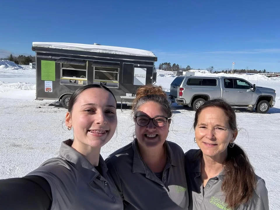 Three women smiling in front of a food truck in a snowy landscape.