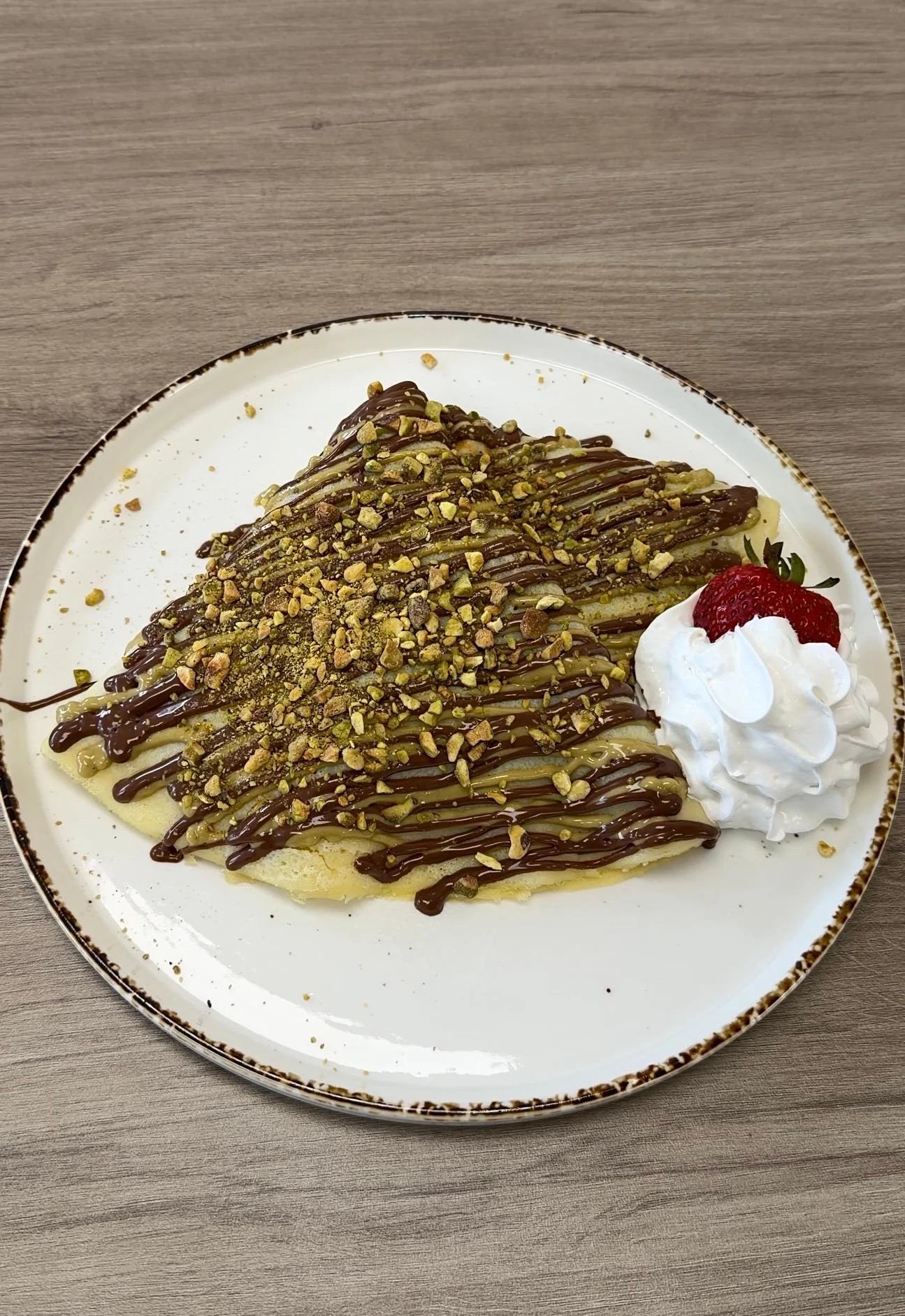 A delicious crepe filled with a sweet spread, topped with crushed pistachios, drizzled chocolate, and a dollop of whipped cream accompanied by a fresh strawberry.