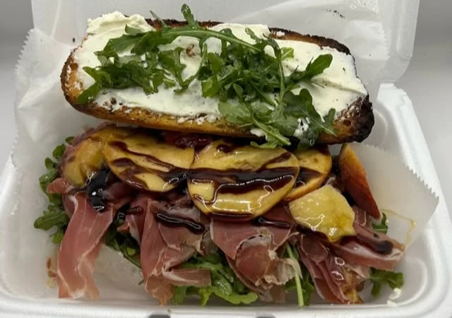 A gourmet sandwich featuring prosciutto, arugula, and a slice of grilled peach drizzled with balsamic glaze.