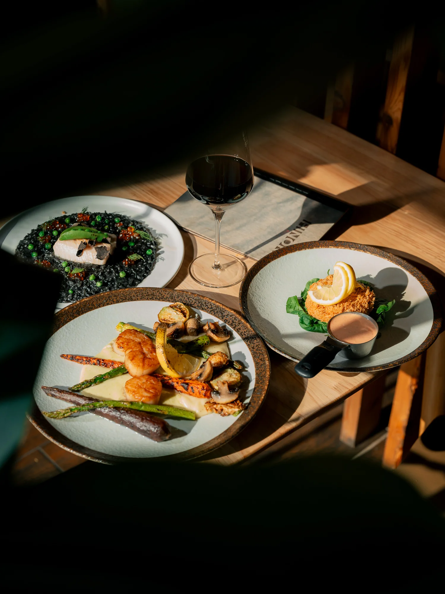 An elegant dining table set with three gourmet dishes, including seafood on a bed of black rice, accompanied by a glass of red wine.