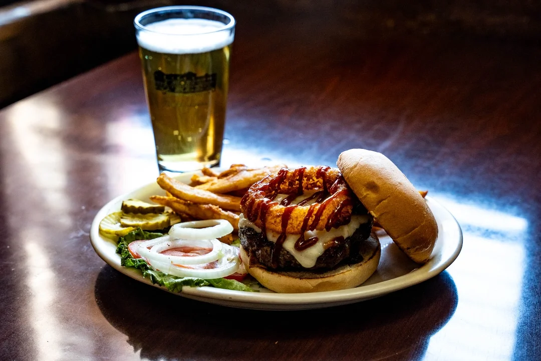 A gourmet burger topped with crispy onions and served with a side of fries, pickles, and lettuce. Accompanied by a glass of beer.