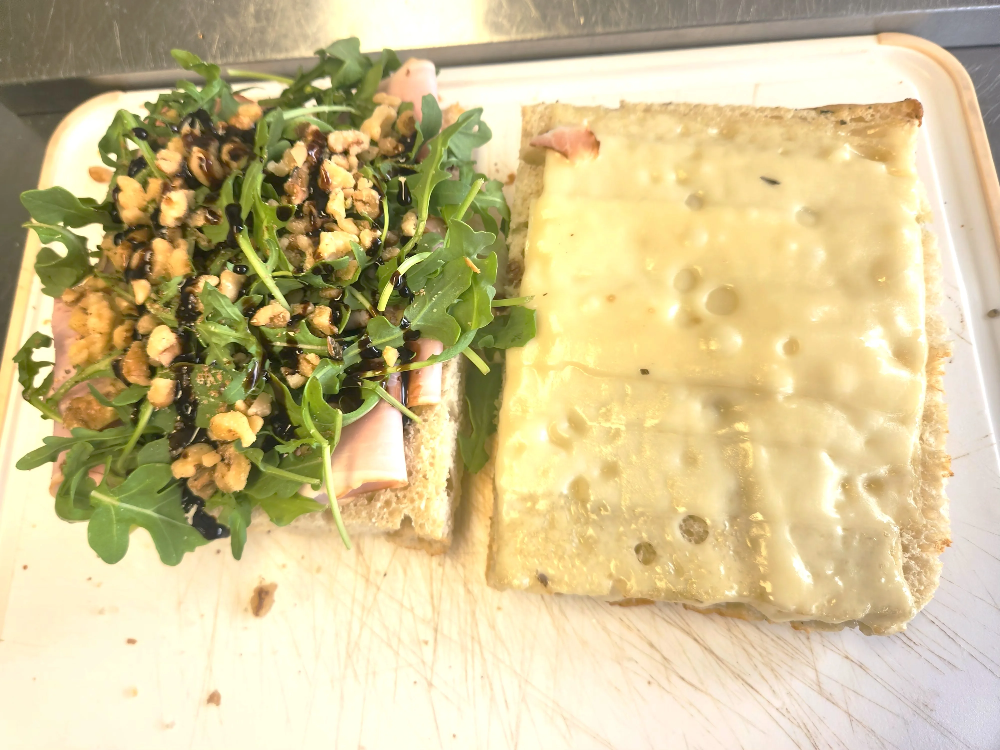 A sandwich with layers of fresh arugula, nuts, and ham on one side, and a slice of melted cheese on the other. The sandwich is served on a white cutting board.