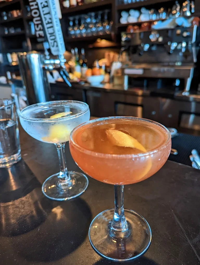 Two elegant cocktails served in stemmed glasses, one garnished with a lemon twist, set against a bar backdrop.