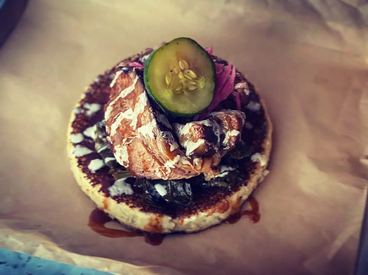 A round corn-based dish topped with meat, greens, pickled onions, and a slice of cucumber.