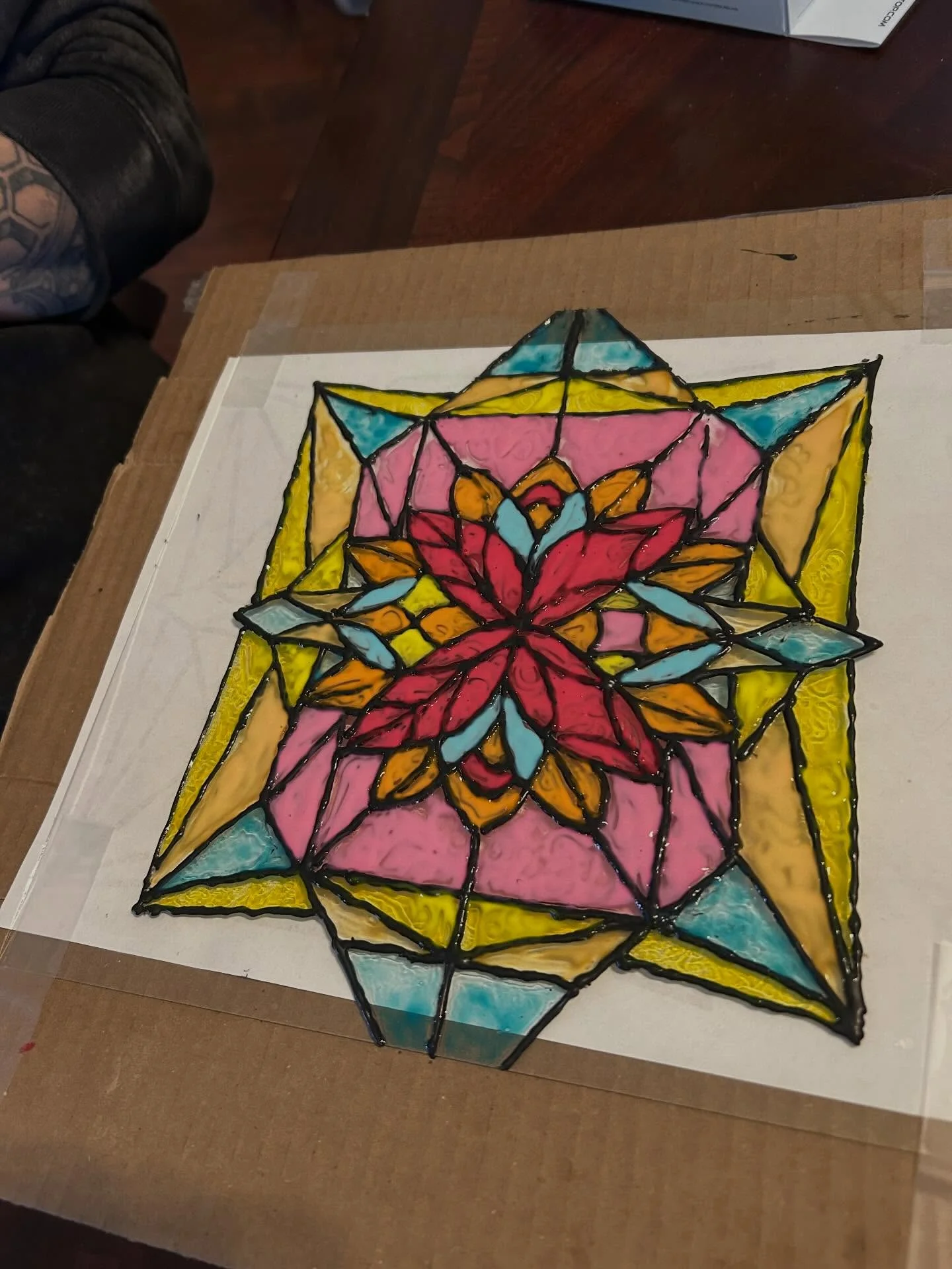 A colorful stained glass design featuring a floral motif, with vibrant shades of red, pink, yellow, and blue. The artwork is outlined with black lines and placed on a cardboard surface.