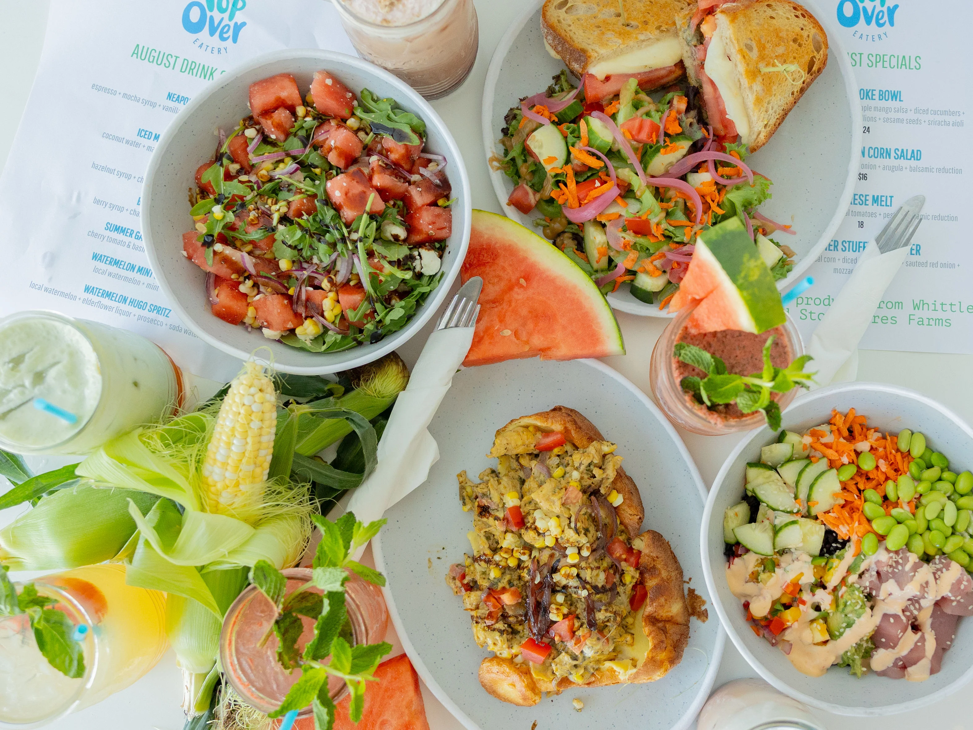 A vibrant spread of fresh salads, sandwiches, and drinks. The table features a watermelon salad, various greens, a sandwich, and colorful beverages along with fresh produce.