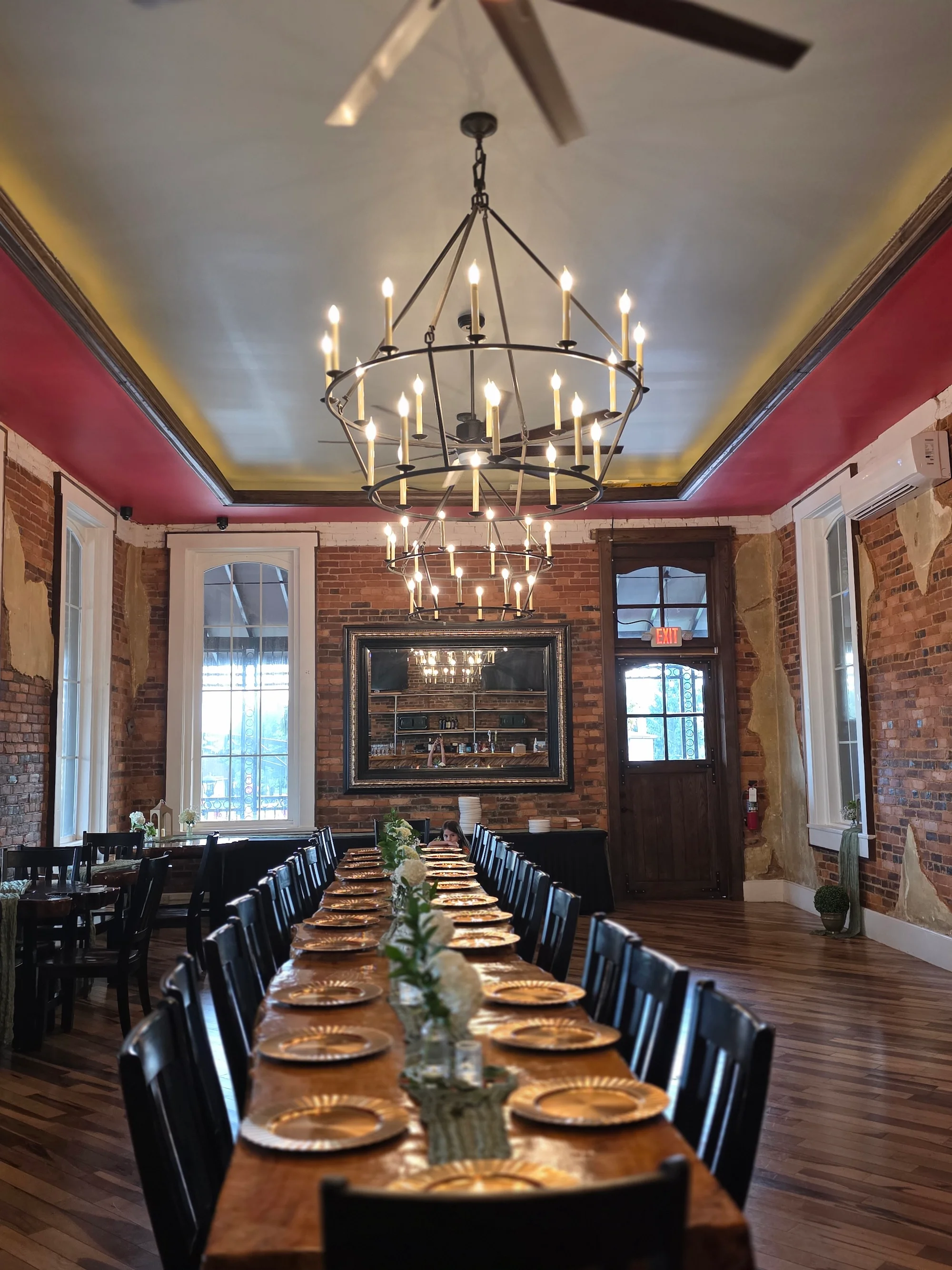 A stylish dining room with a long wooden table set for an event, featuring an elegant chandelier and exposed brick walls.