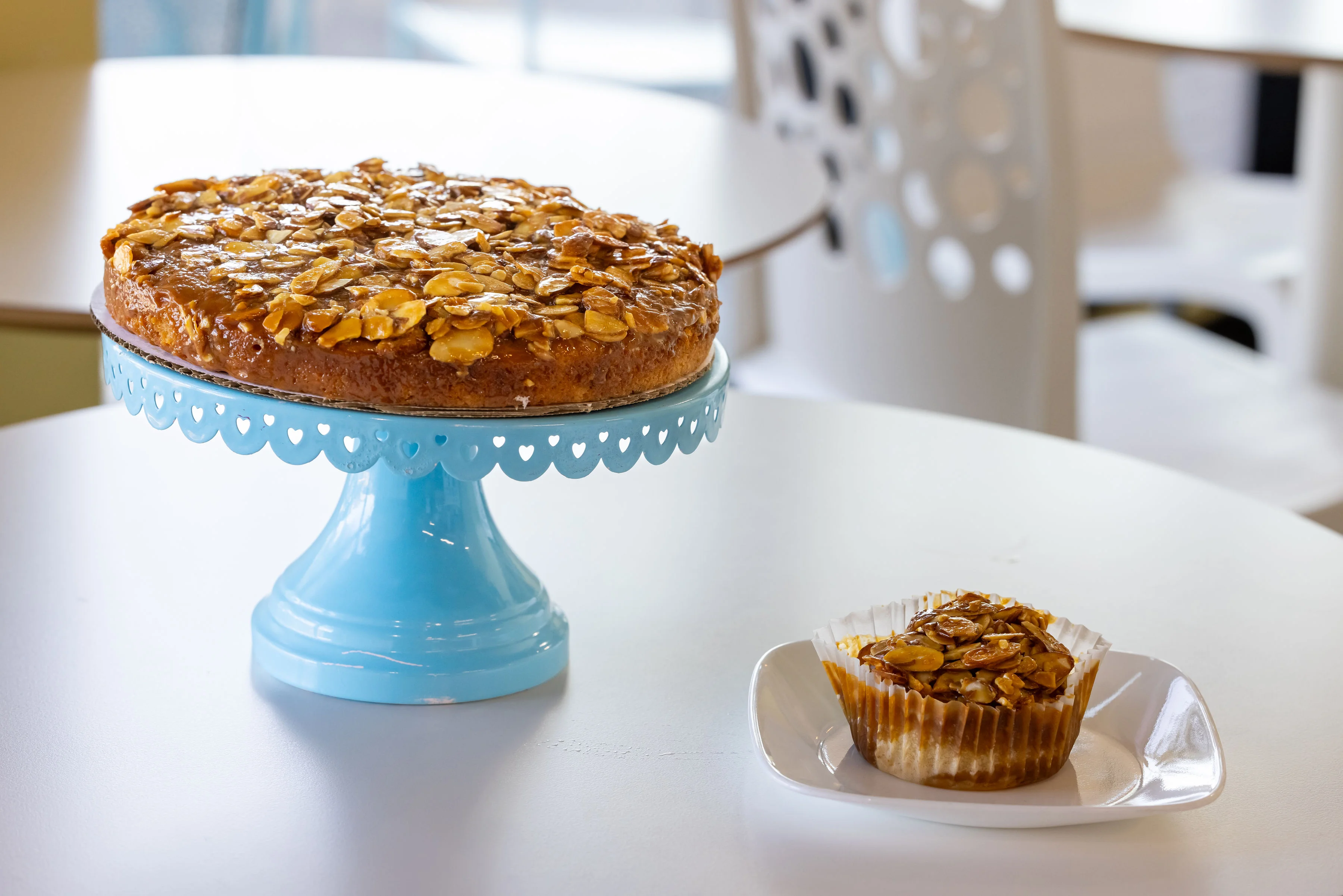 A round cake topped with almond slices on a blue cake stand, accompanied by a similar almond muffin on a white plate.