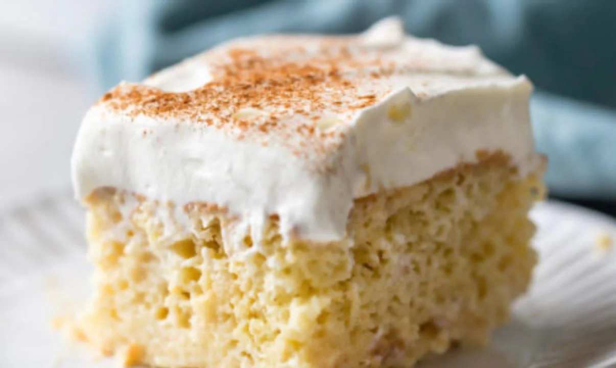 A slice of Tres Leches cake on a white plate, showcasing its moist texture from being soaked in three types of milk. The cake is topped with whipped cream and a sprinkle of cinnamon, served on a wooden table.