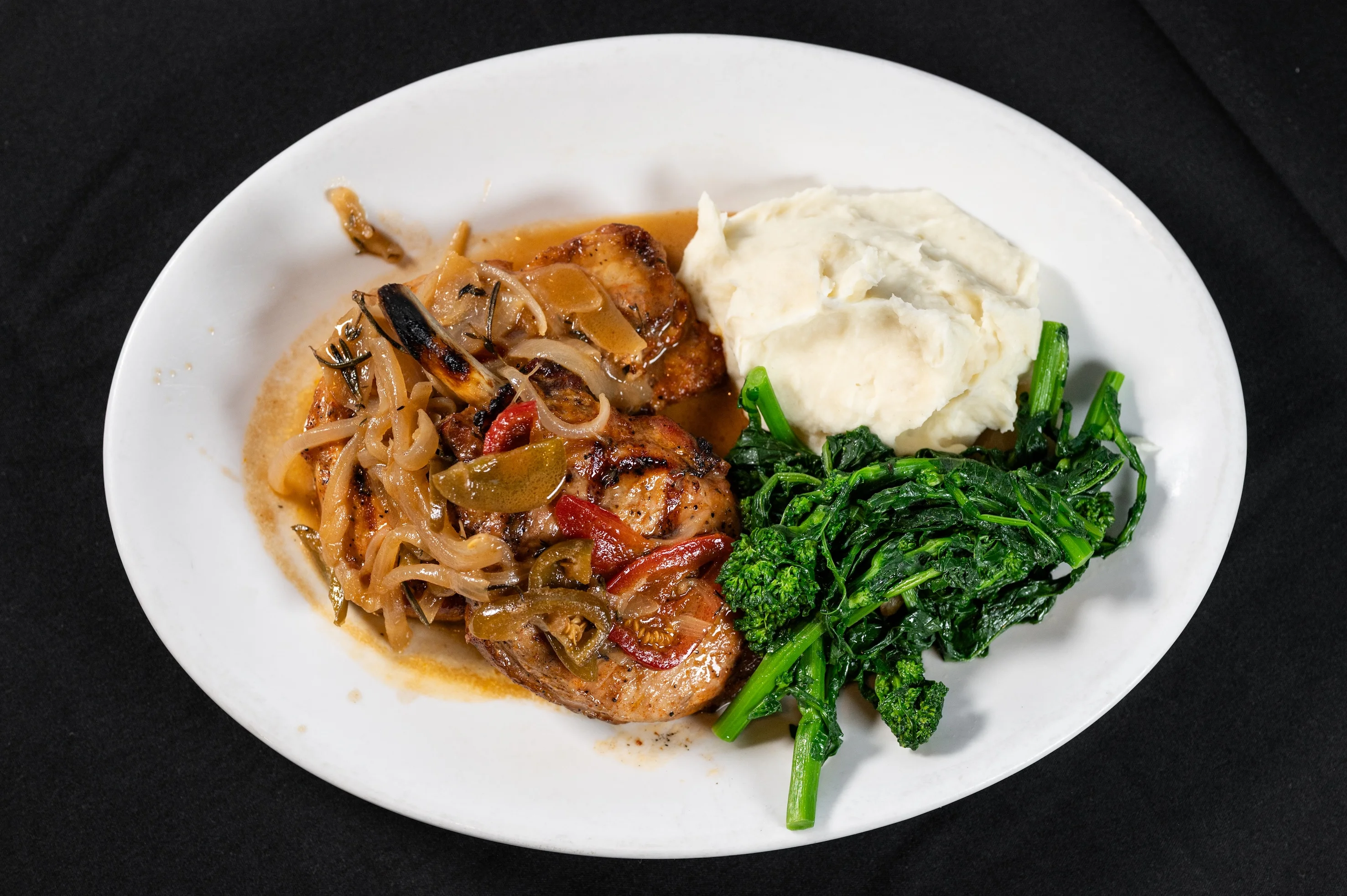 A white plate with juicy pork chops topped with sautéed onions and peppers, accompanied by creamy mashed potatoes and vibrant sautéed greens served at Toscana on Main Eastchester NY