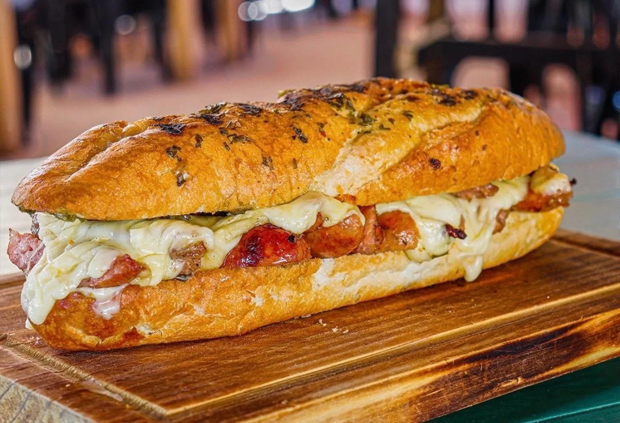 A long sandwich filled with grilled meats and melted cheese, served on a wooden board.
