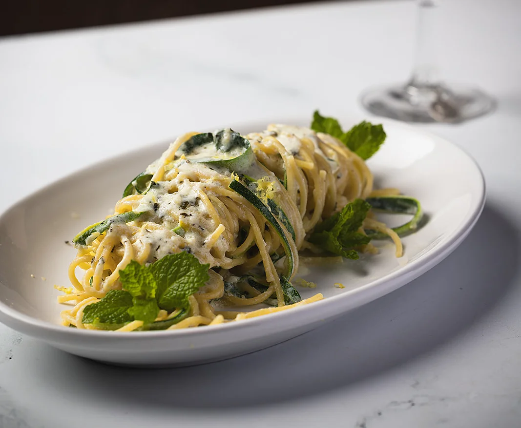 A serving of spaghetti with creamy sauce and fresh mint leaves, accompanied by a cocktail in the background.