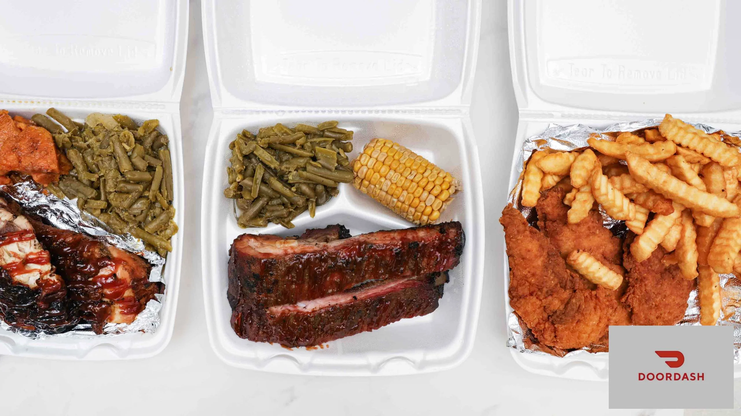 Three takeout containers with a variety of dishes including ribs, fried chicken, green beans, corn, and fries, showcasing a hearty meal.