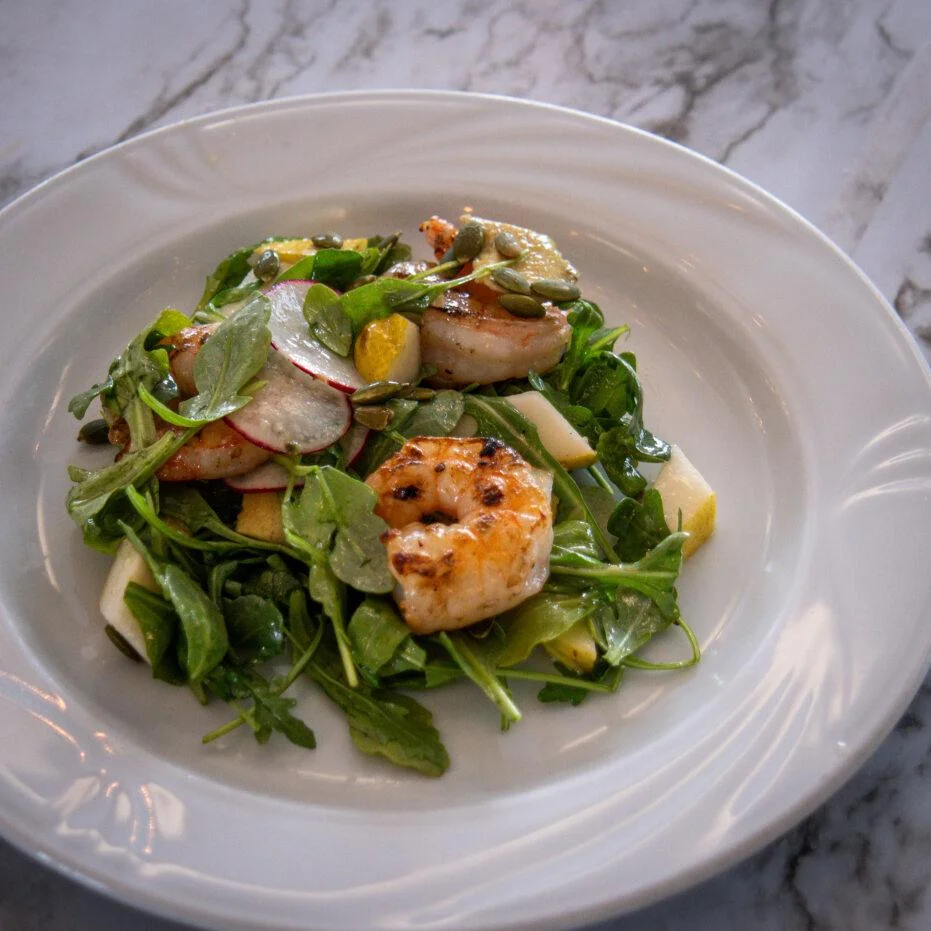 A fresh salad with grilled shrimp, mixed greens, and various toppings including radishes and pumpkin seeds served in a white bowl.