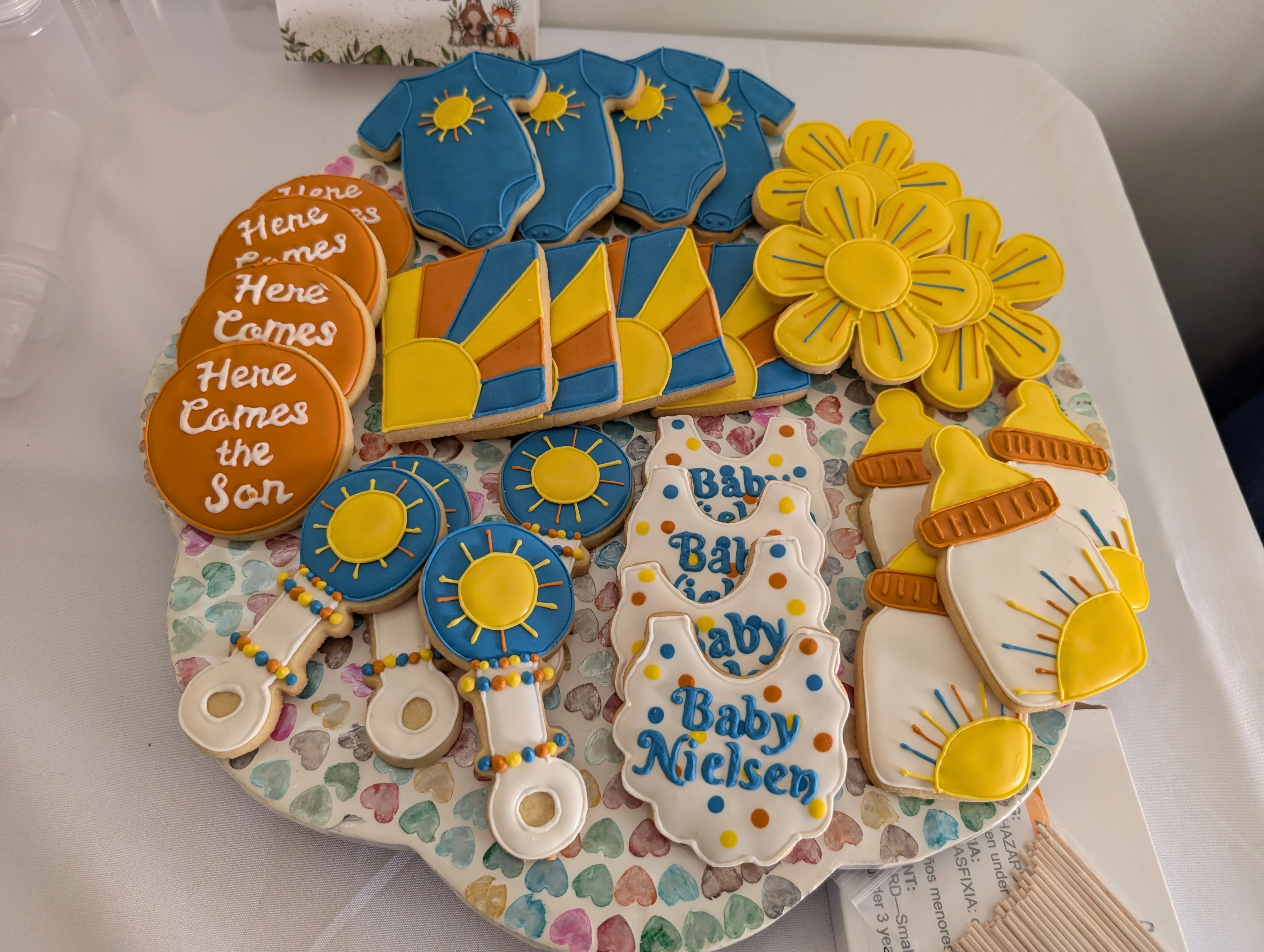 A platter of baby shower cookies in muted orange, blue, yellow, and white with a "Here Comes the Son" theme with sun onesies, rattles, and bottles.