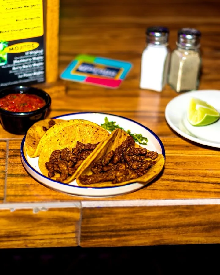 A plate of tacos filled with seasoned meat, garnished with onions and fresh herbs. Accompanied by a dish of salsa and a lime wedge.