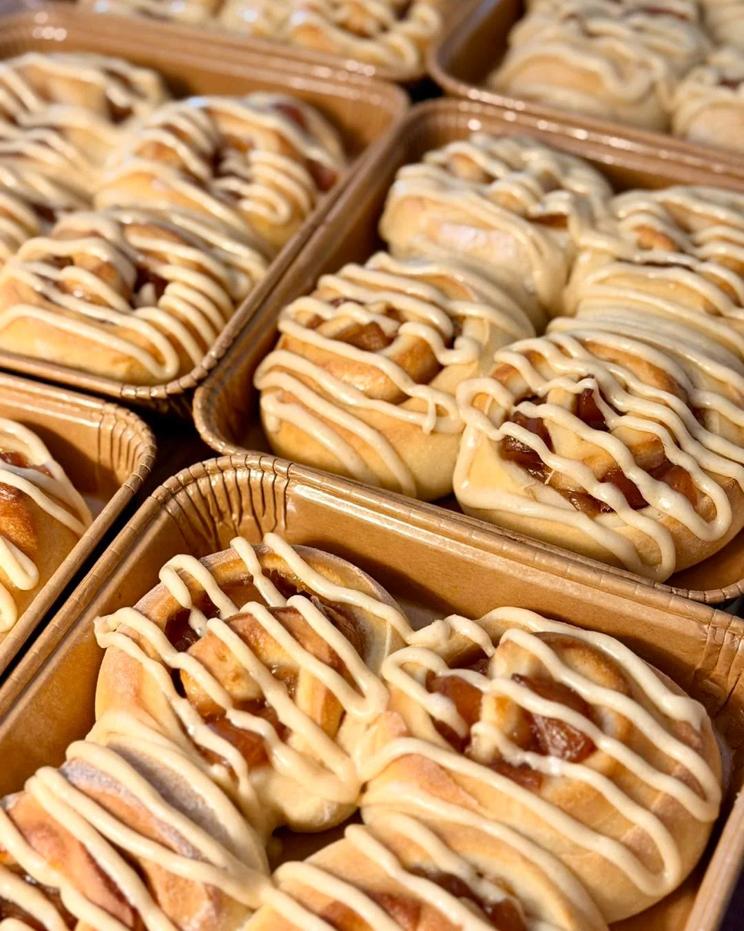 Freshly baked sweet rolls drizzled with icing, arranged neatly in trays.