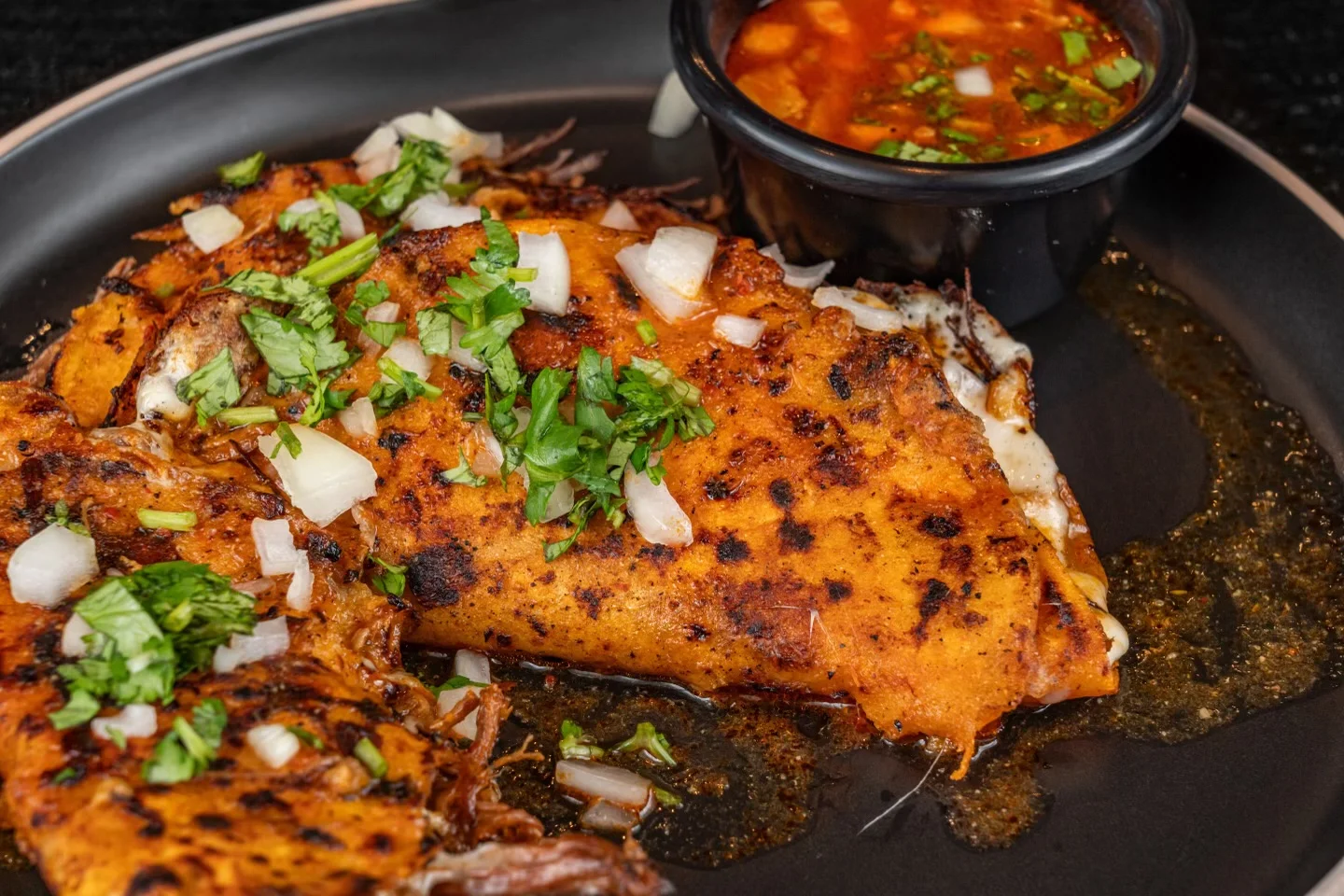 A plate of grilled enchiladas topped with chopped onions and cilantro, served with a side of red sauce.