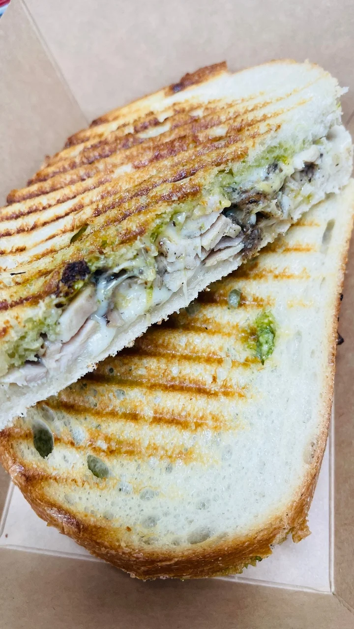 A toasted sandwich cut in half, revealing chunks of chicken and a green spread inside, served in a cardboard box.