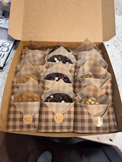 A box filled with neatly arranged cookies, each placed in a decorative paper wrapper. The cookies appear golden brown and chocolate chip.