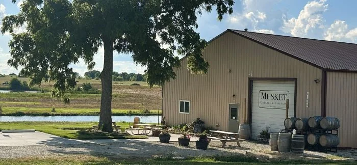 Exterior view of Musket winery with seating and barrels by a pond.