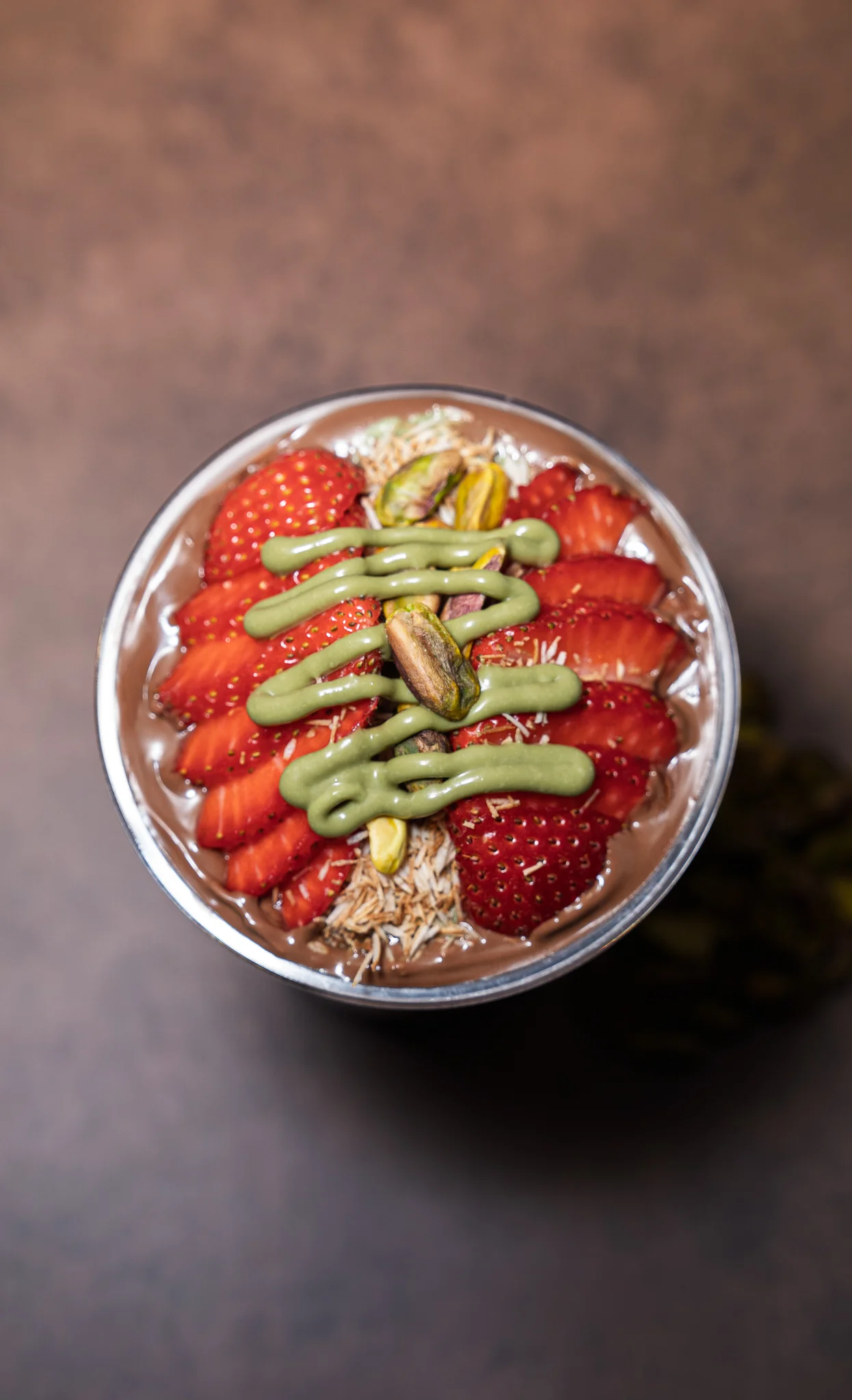 A bowl filled with a creamy dessert topped with strawberries, pistachios, and a green sauce, garnished with shredded coconut.