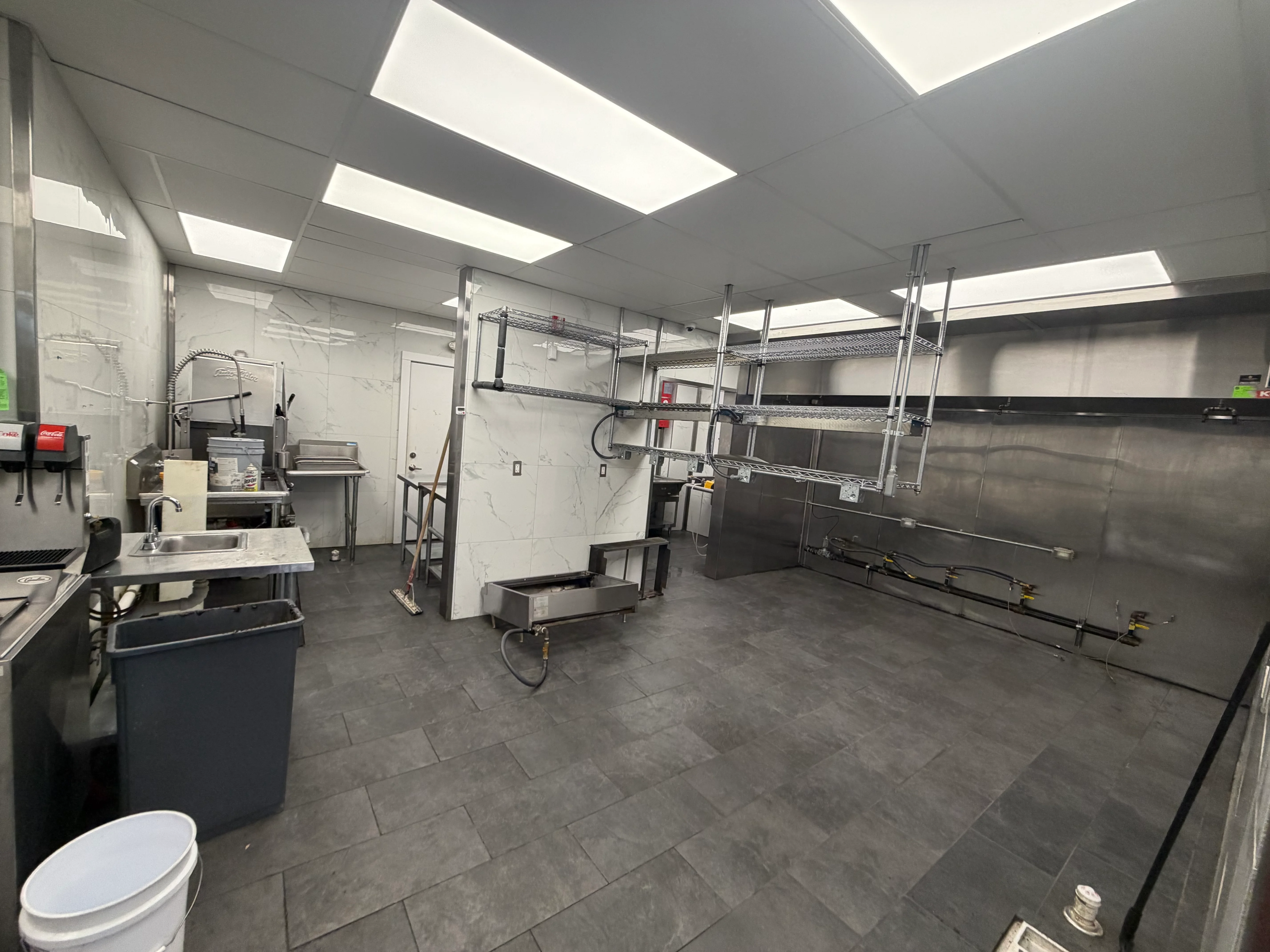 A commercial kitchen space with stainless steel fixtures, a large sink, and a clean, minimalist design. The flooring is dark tile, and bright overhead lights illuminate the area.