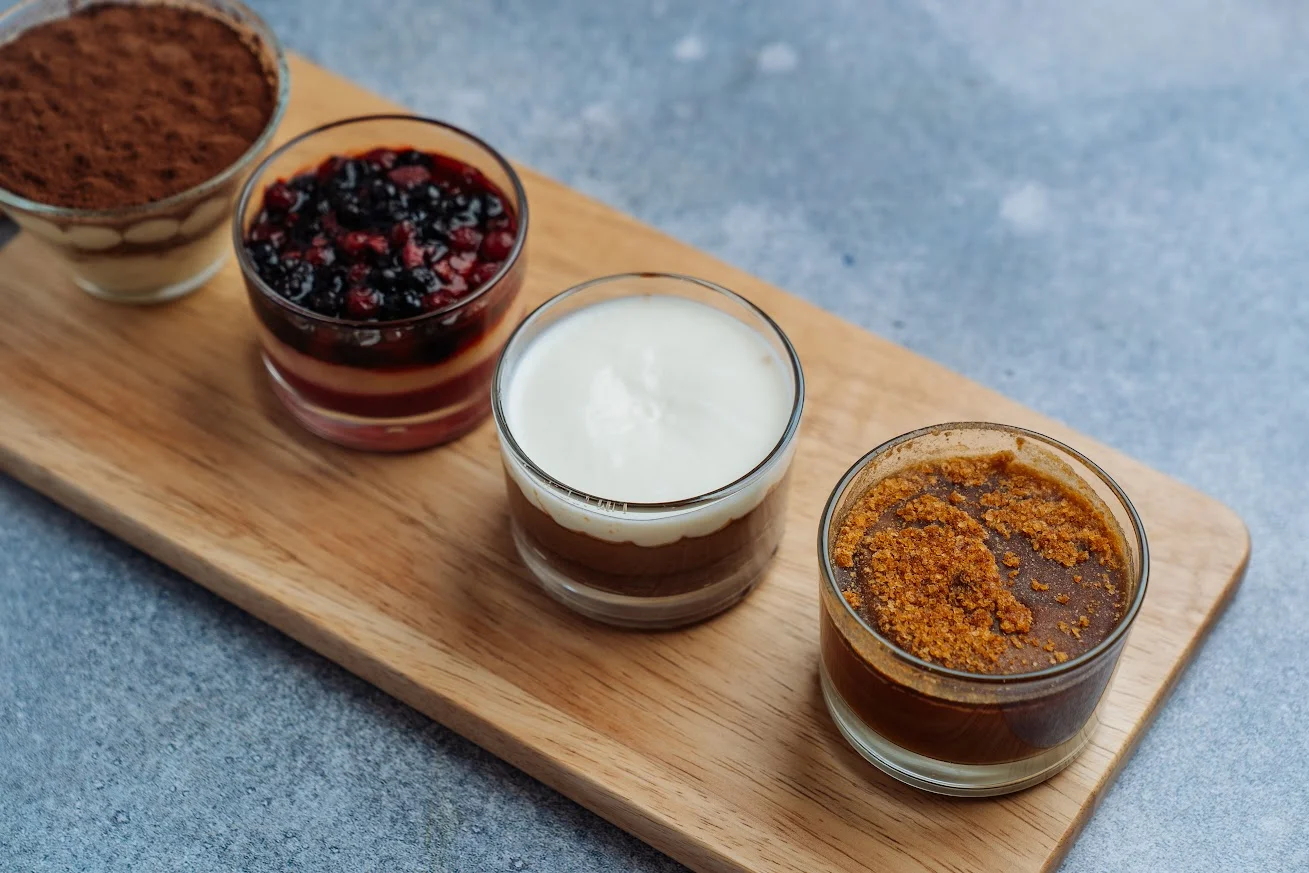A variety of small dessert cups displayed on a wooden board, featuring layered sweets, fruits, and creamy textures.