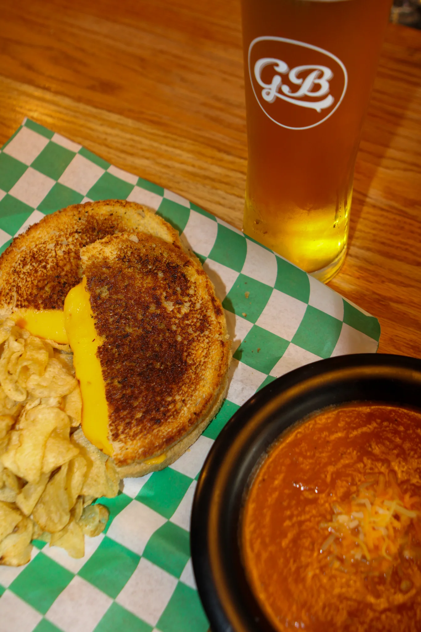 A grilled cheese sandwich with melted cheese, served with crispy chips and a bowl of creamy tomato soup. A cold beverage is also visible in the background.