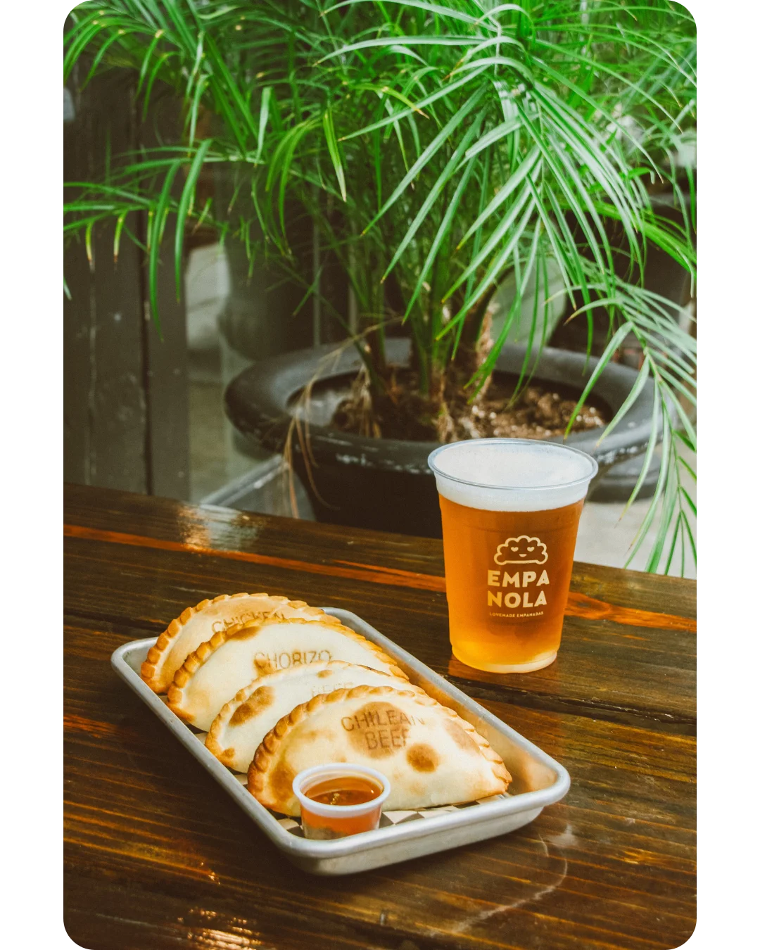 A plate featuring several empanadas labeled 'CHORIZO' and 'CHILEAN BEEF', accompanied by a cold beverage and dipping sauce, with greenery in the background.