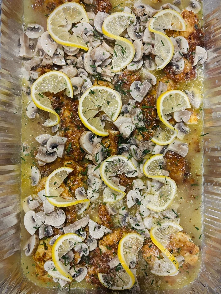 A tray of chicken topped with lemon slices and mushrooms, garnished with herbs.