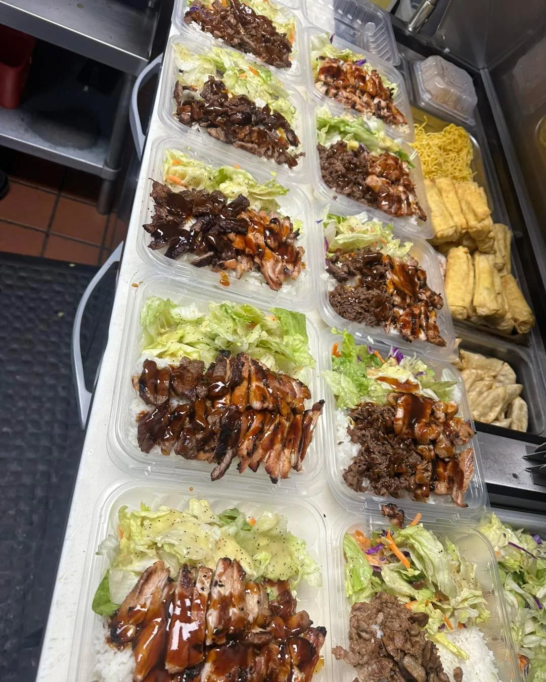 A row of meal containers filled with rice, protein, and fresh vegetables, all prepared for serving.