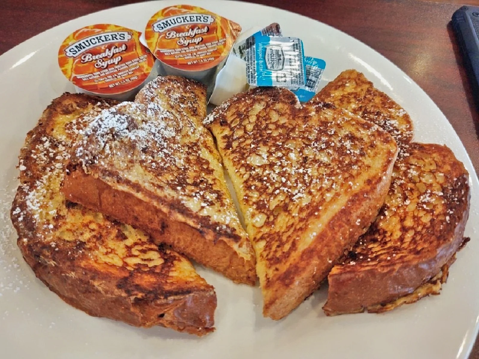 A plate of golden French toast dusted with powdered sugar, served with packets of breakfast syrup and whipped butter on the side.