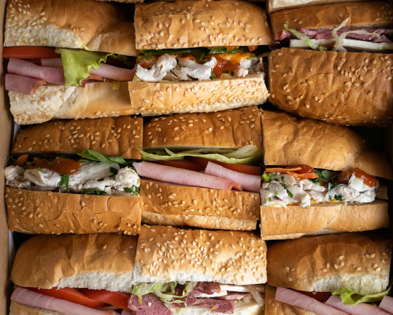 Overhead view of assorted sub sandwiches on sesame and plain hoagie rolls, filled with deli meats, lettuce, tomato, and chicken salad. The sandwiches are tightly packed together in a catering-style arrangement