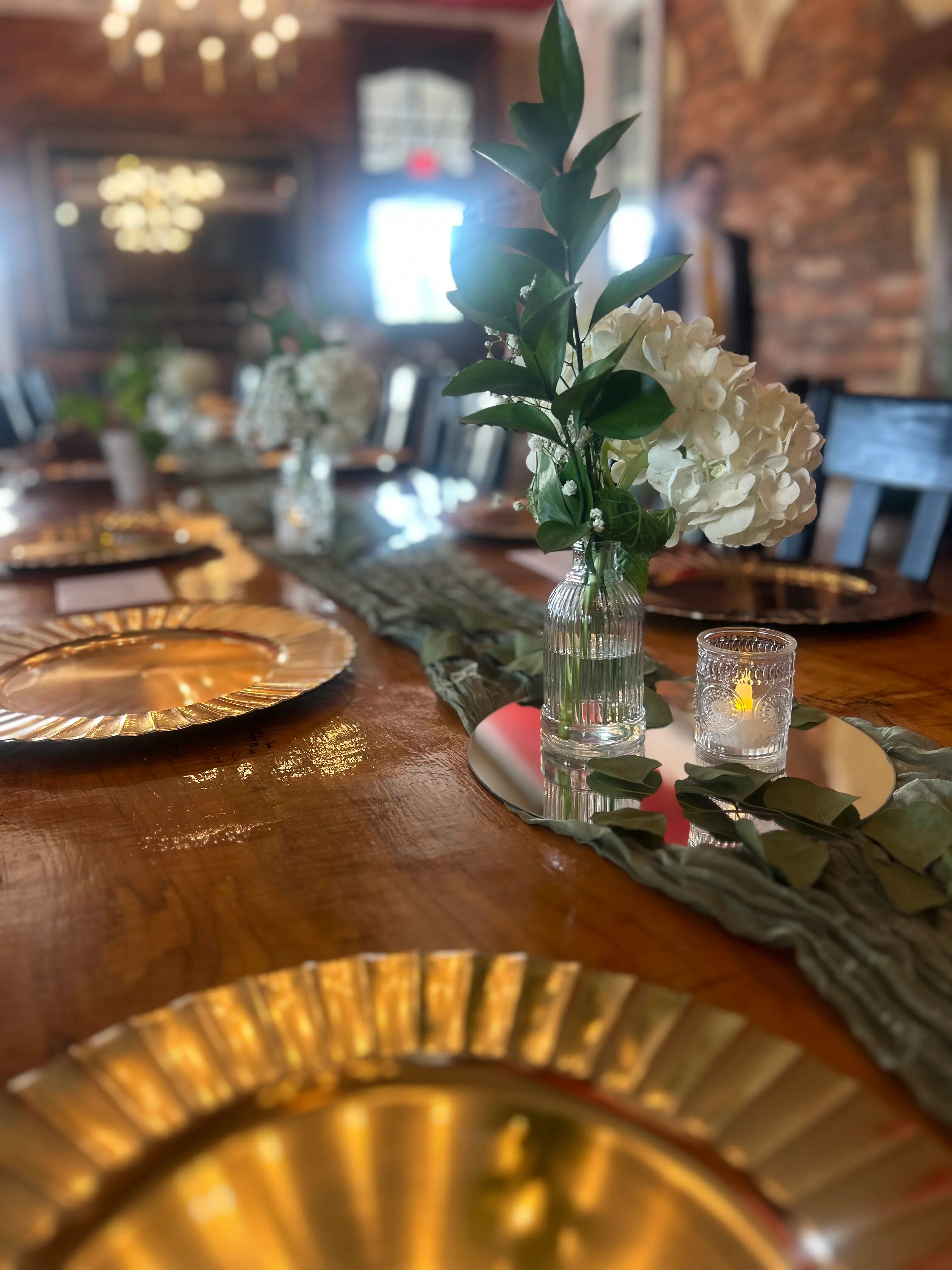 A beautifully arranged dining table featuring golden plates, greenery, and floral centerpieces, set against a rustic brick wall.