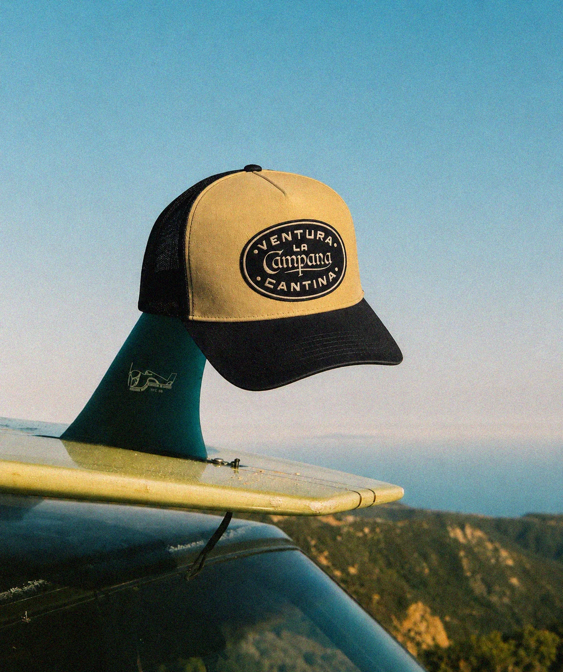 A cap resting on the fin of a surfboard, positioned on a car roof. The background features a clear sky and distant hills.