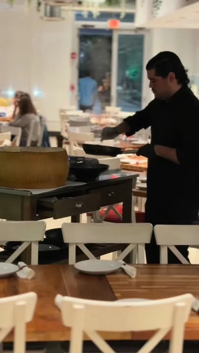 A chef preparing food in a bustling restaurant, with tables and chairs in the background.