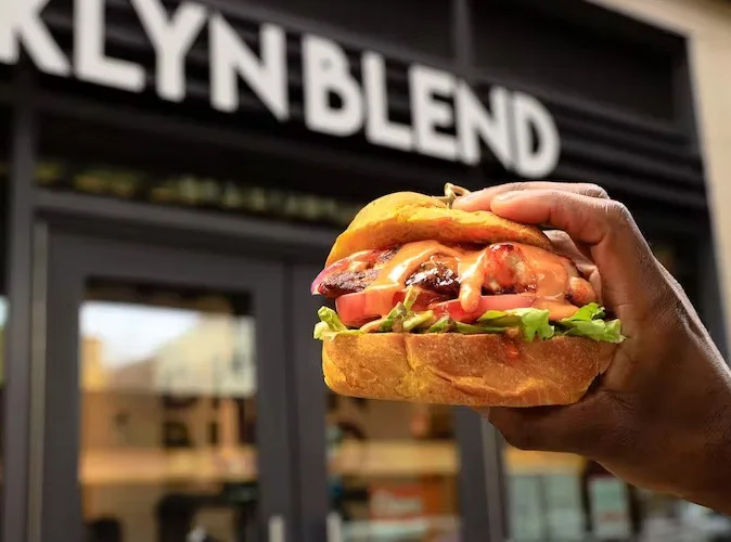 Close-up of a hand holding a juicy burger with lettuce, tomato, and sauce oozing from the bun. The blurred background shows the exterior of a building.