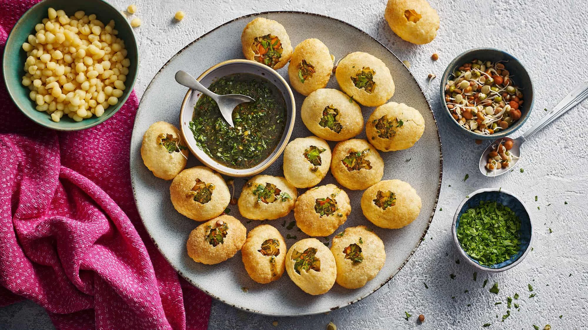 A platter of crispy golden puris served with a green dipping sauce and various toppings, including lentils and sprouts, arranged on a decorative plate.