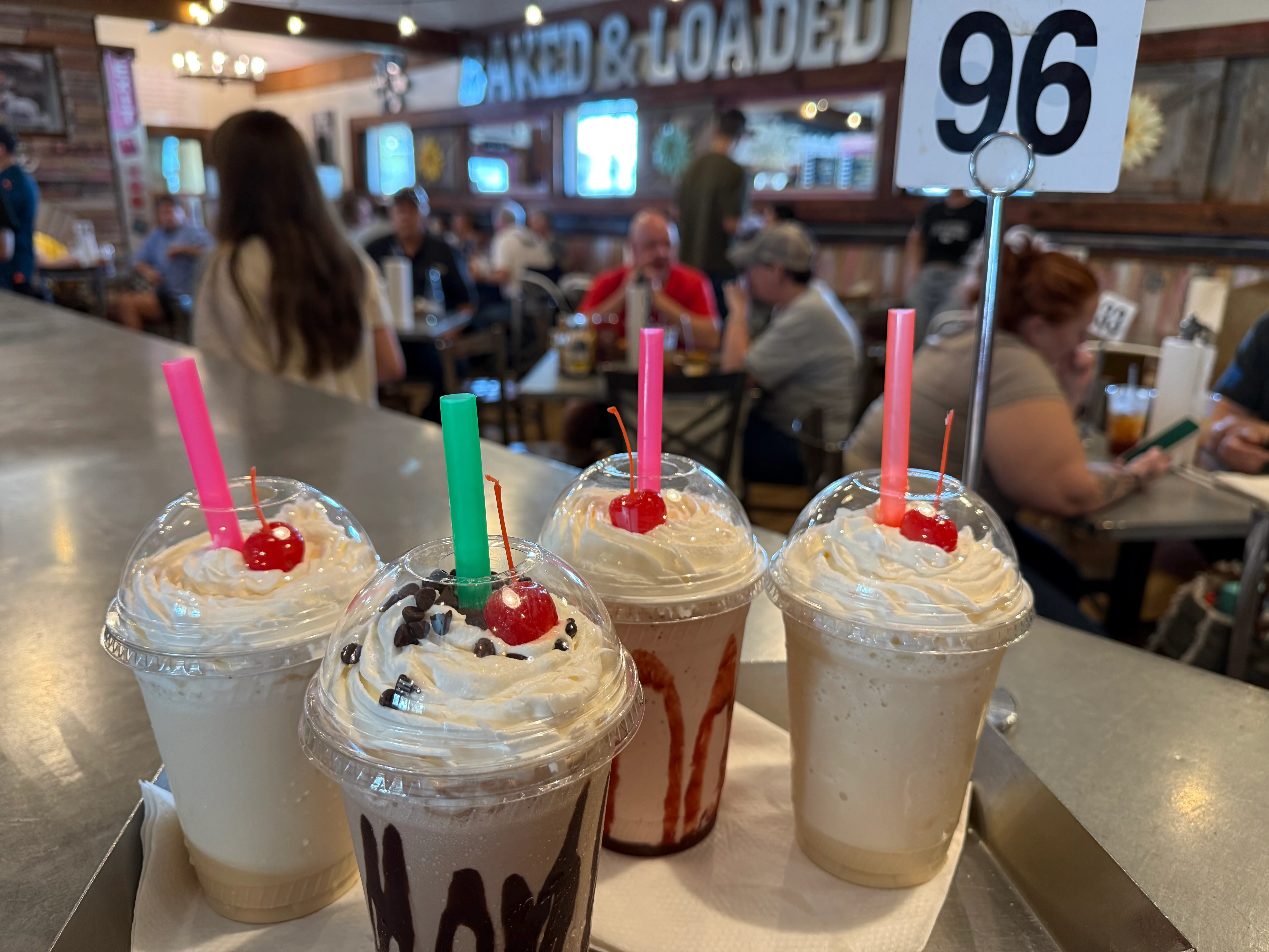 Four milkshakes topped with whipped cream, cherries, and colorful straws served on a tray in a busy restaurant.