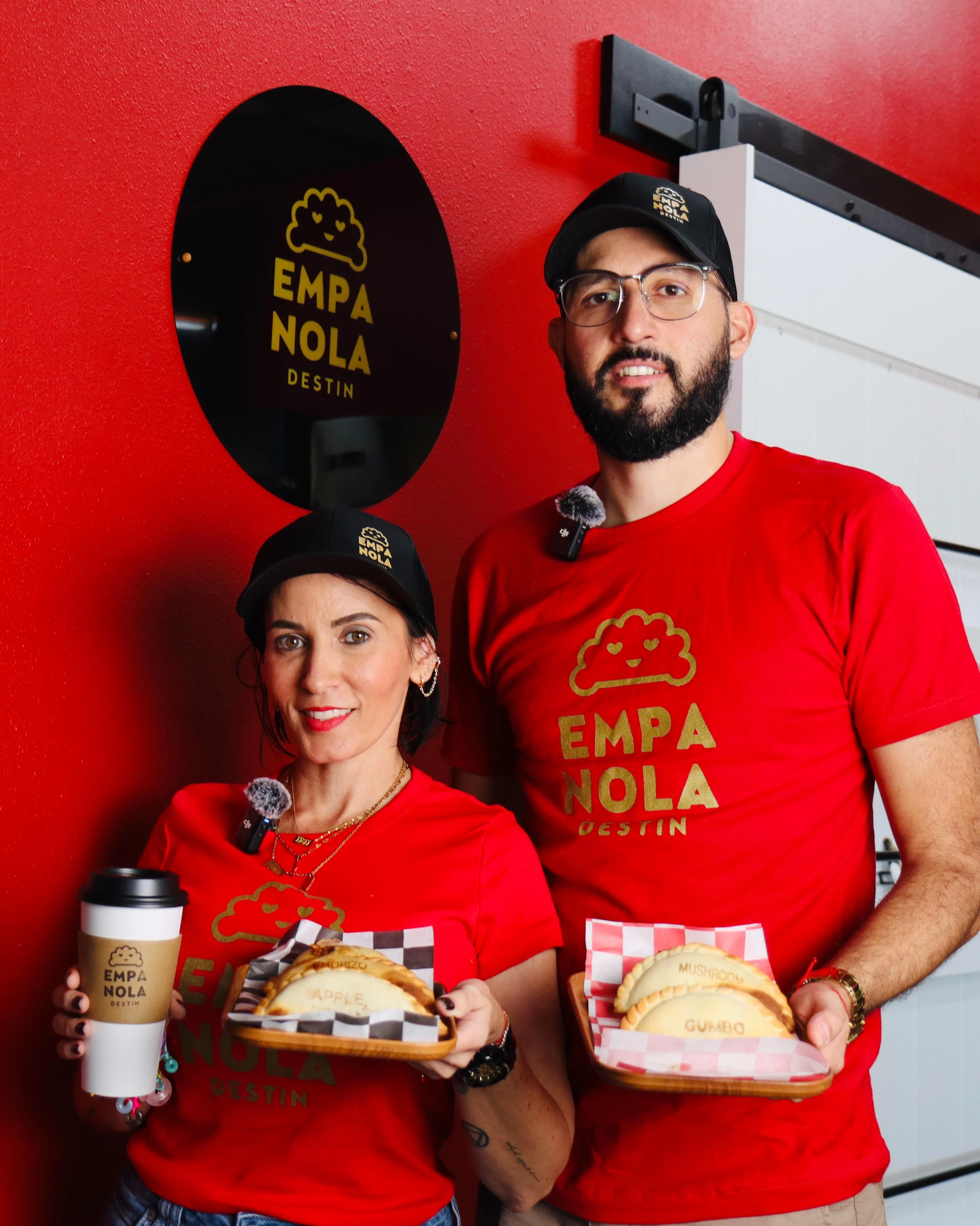 Two individuals holding empanadas in a cozy restaurant setting decorated with a bold red wall and the restaurant's logo.
