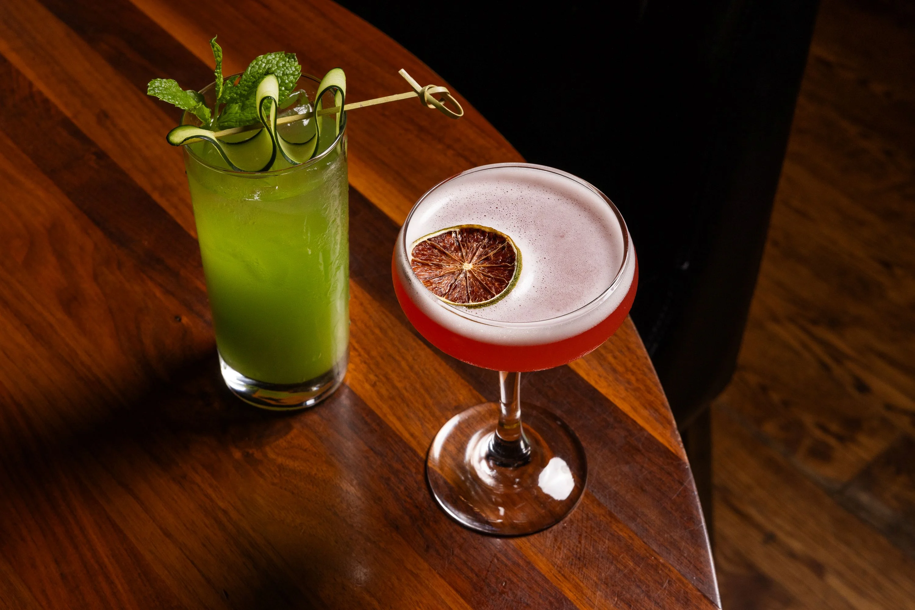 Two colorful cocktails on a wooden table, one green and one pink.