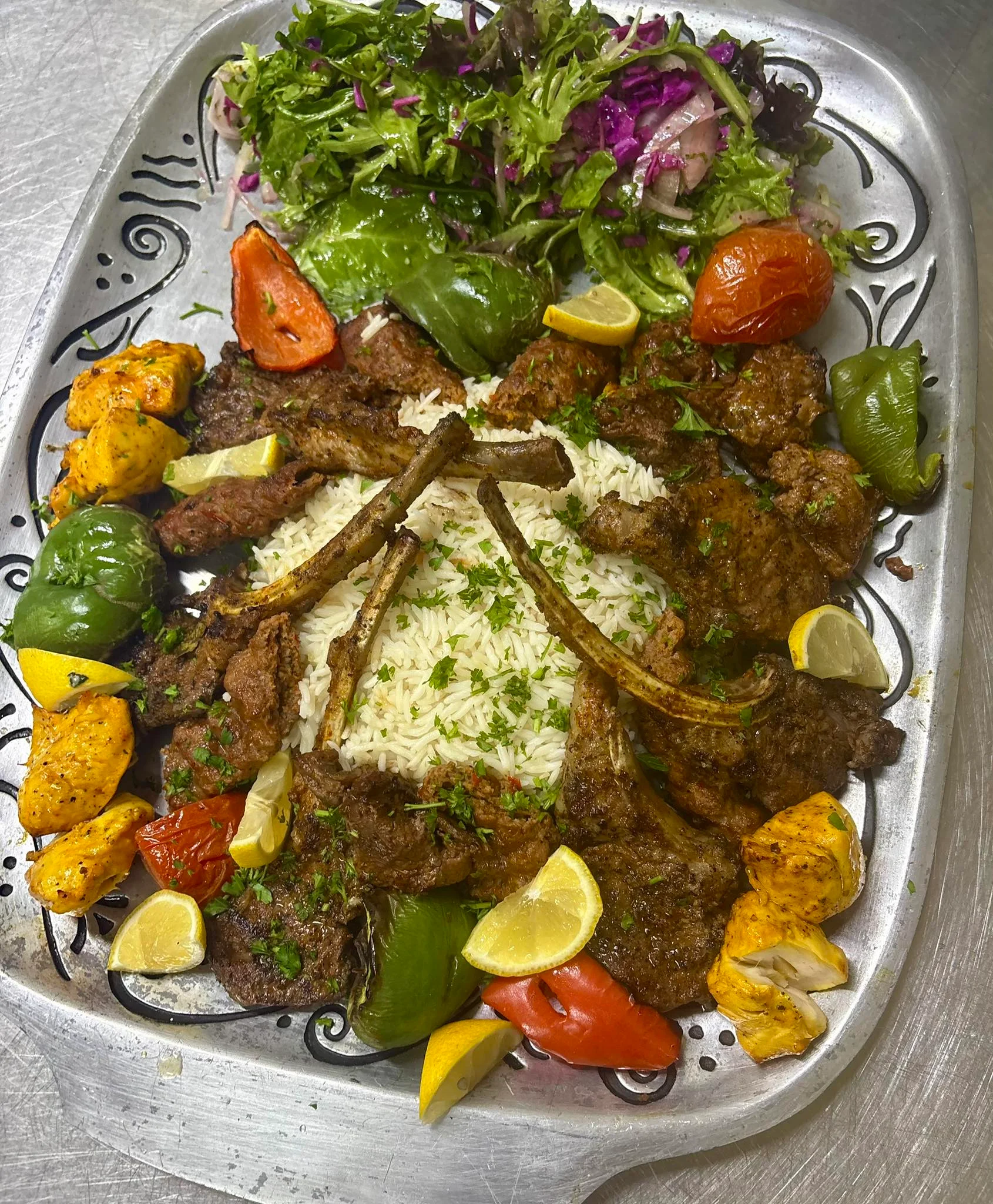 A platter showcasing a variety of grilled meats, rice, and colorful vegetables, garnished with lemon and herbs on a silver serving tray.