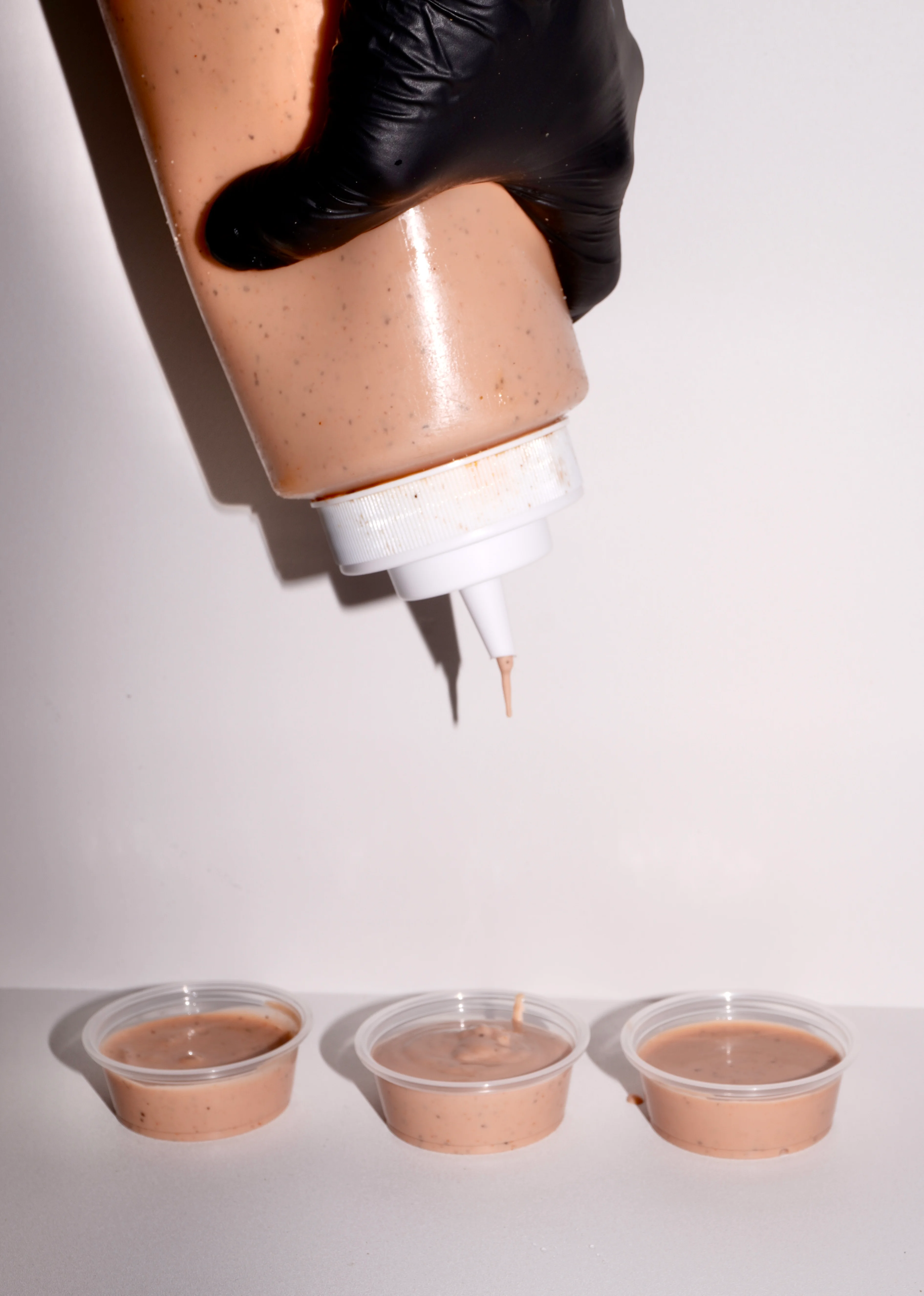 A hand in a black glove holds a squeeze bottle filled with a pink sauce, positioned above three small clear containers filled with the same sauce.